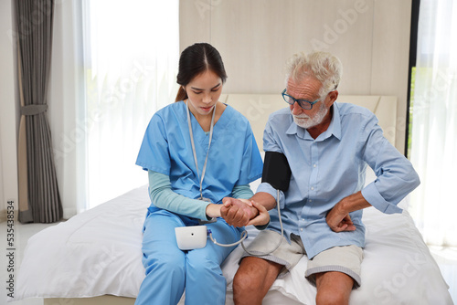 Happy caregiver checking blood pressure monitor and heart rate senior elderly man patient on bed at retirement house. Asian smiling nurse taking good help care and support of elder patient at house