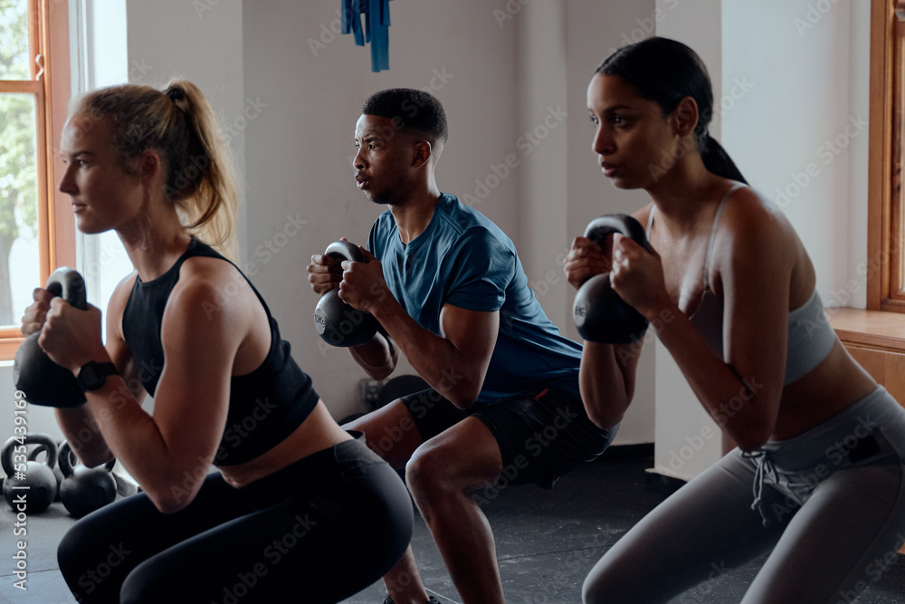 Naklejka premium Three focused multiracial young adults doing kettlebell squats at the gym