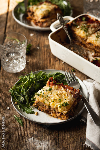 Baked Vegan lasagna casserole with eggplant and cashews on a rustic wooden table served for two on plates with some arugula  and a lot of vegan shredded cheese