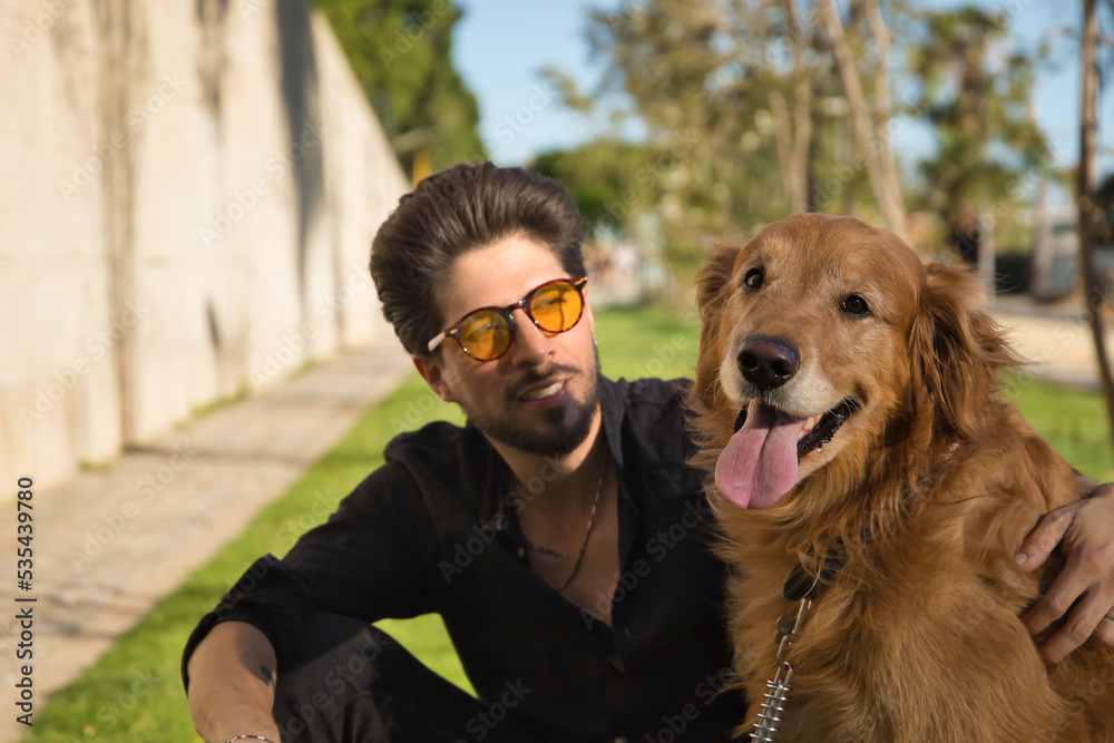 Portrait of a young Hispanic man with a beard, sunglasses and black shirt, sitting on the lawn next to his dog sunbathing. Concept animals, dogs, love, pets, gold. Selective focus on the dog.