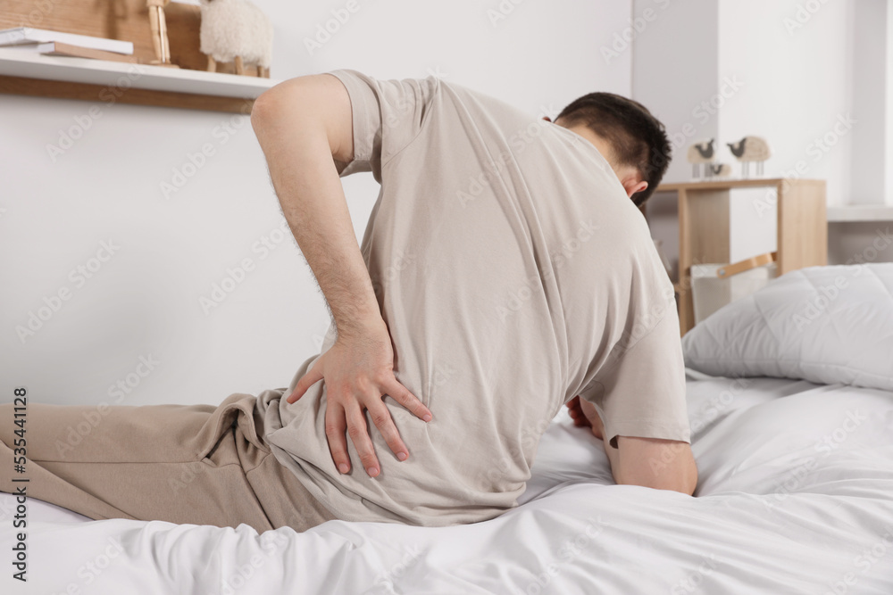 Man suffering from back pain while sitting on bed in room. Symptom of scoliosis Man suffering from back pain while sitting on bed in room. Symptom of scoliosis