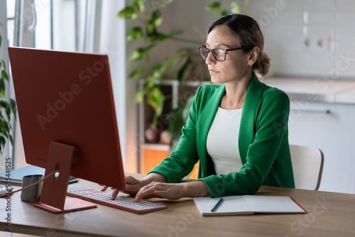 Successful focused Caucasian businesswoman working on computer sits at office desk. Business lady in glasses and green jacket is typing order for company employees or e-mail letter for partners