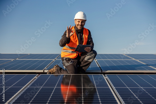 The worker is crouching on the rooftop with solar panels with tablet in his hands and approving. The solar panels passed the test.