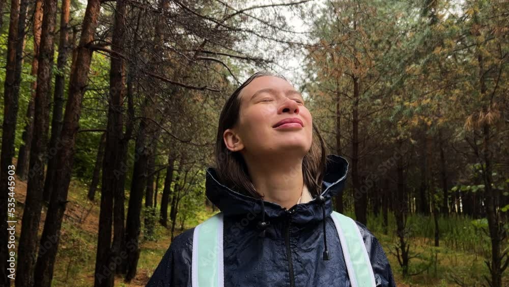 Young woman in blue raincoat stands under the rain in forest, looks up, inhale and breathe in fresh air. Girl with closed eyes, wet hair portrait. enjoy the rain drops to her face smiling. autumn mood