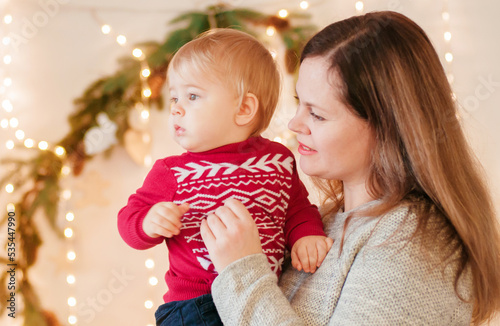 A young blond-haired mother with her little son in her arms in a room decorated with pine needles and sparkling garlands for Christmas. Christmas mood
