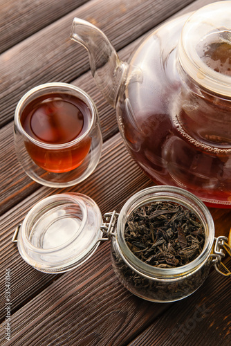 Cup and teapot of black tea and dried leaves on wooden background, closeup