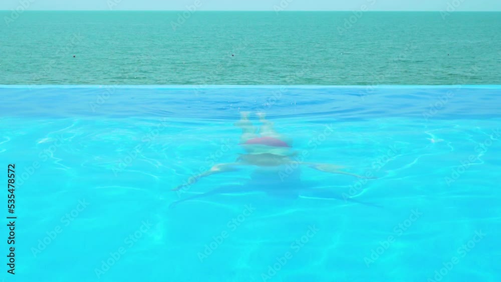 Vacation Woman Swims Slowly Underwater in Outdoor Infinity Swimming Pool of Luxury Resort on Summer Sunny Day. Concept of Luxurious Comfortable Holidays. Vacation Woman Swims Slowly Underwater in Outdoor Infinity Swimming Pool of Luxury Resort on Summer Sunny Day. Concept of Luxurious Comfortable Holidays.
