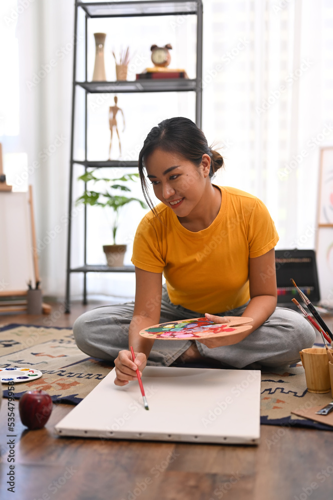 Millennial beautiful woman painting picture on canvas with oil paints in bright home studio. Leisure activity and art concept