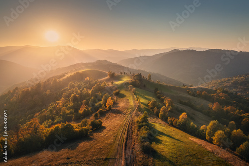 Fototapeta Naklejka Na Ścianę i Meble -  Aerial view of beautiful hills, forest and mountains in fog at sunset in autumn in Ukraine. Colorful landscape with woods in fog, meadows, golden sunlight, trees in fall. Nature. Top view from drone
