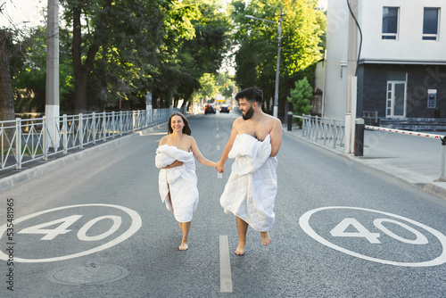 Couple in love, a man and a woman, in white blankets are running along the road in blankets at dawn.