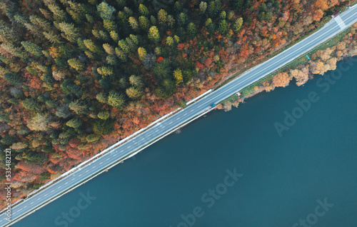 Aerial view of road near blue lake, forest at sunset in autumn. Travel in Ukraine. Top view of beautiful road, orange trees in fall. Colorful landscape with highway and river shore. Road trip. Nature