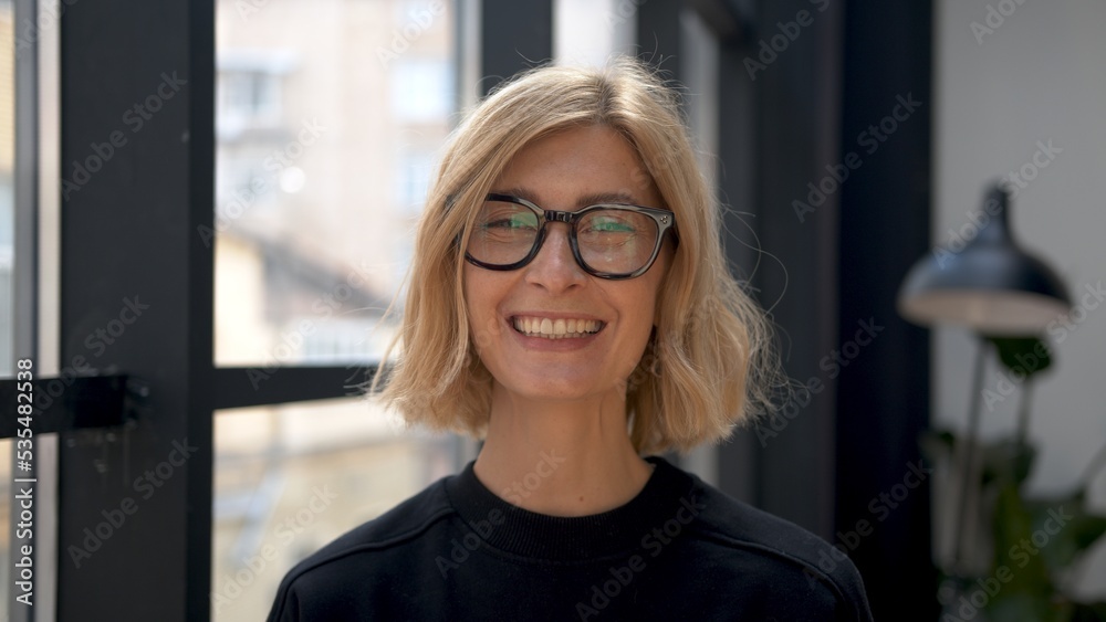 Boss woman smiling in eyeglasses, attractive blonde manager portrait on background of windows. Office room and worker in black clothes. Concept of business partner. 