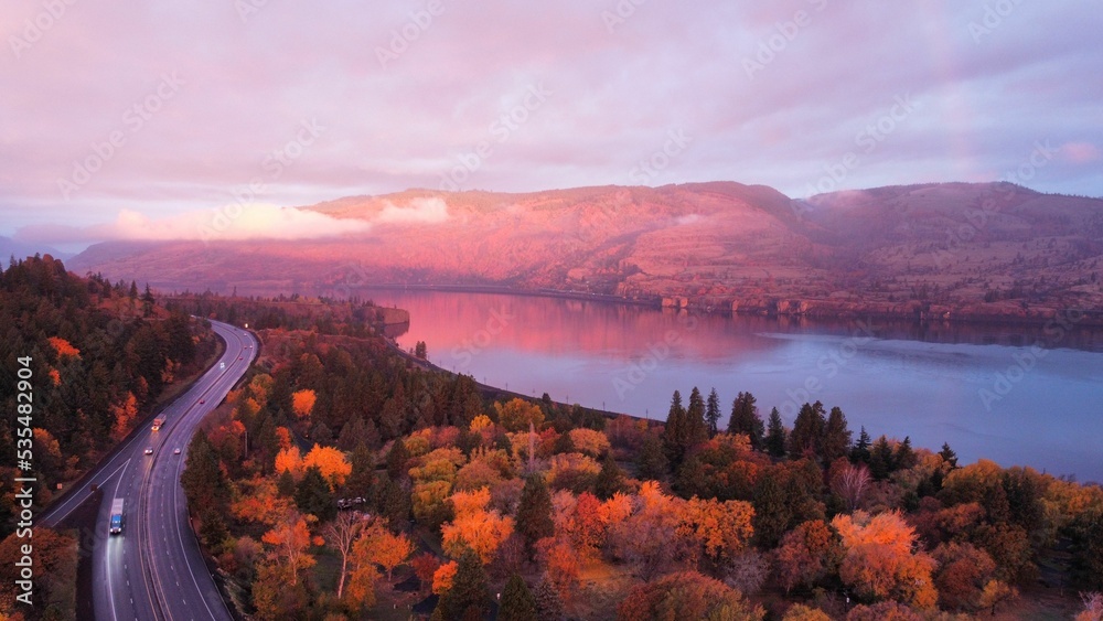 Naklejka premium Aerial view of a lake surrounded by trees and hills near a highway in the Dalles, Oregon at sunset