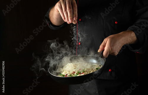 Fototapeta Naklejka Na Ścianę i Meble -  The chef adds salt to the spaghetti in a steaming hot pan. Recipe or menu for restaurant or hotel on black background