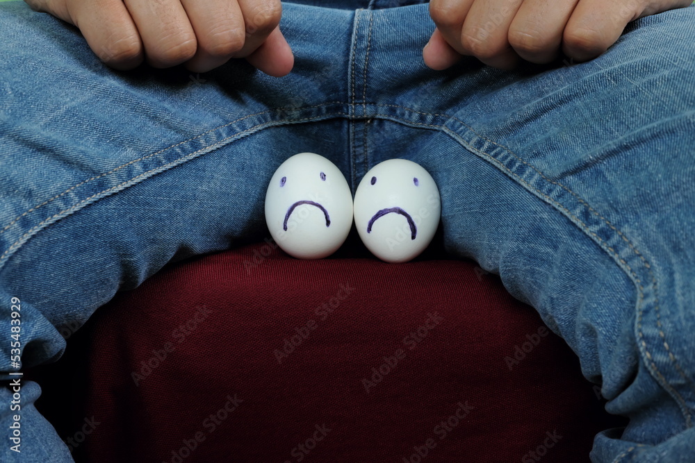 Young Asian man holding two chicken eggs with sad face on crotch groin area. Male infertility, testicular cancer and impotence concept. Young Asian man holding two chicken eggs with sad face on crotch groin area. Male infertility, testicular cancer and impotence concept.