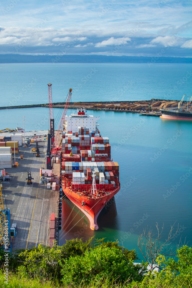 Aerial view over bright red container ship unloading at port in Napier, Hawkes Bay, New Zealand - obrazy, fototapety, plakaty 