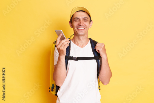 Horizontal shot of smiling optimistic delivery man in yellow cap standing with thermal backpack holding mobile phone, using gadget for his work isolated on yellow background.