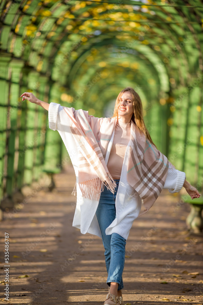 Naklejka premium Beautiful happy young woman with light brown hair walks along a picturesque alley in an autumn park. An attractive woman wrapped herself in a plaid. Fall season. Amazing autumn mood.