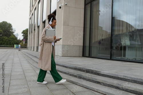 an african american female student in headphones and a laptop in her hands goes to the office building, Concept of a successful marketing strategy