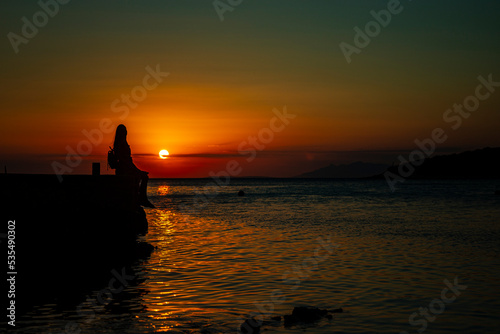 dark silhouette of a beautiful long-haired girl sitting by the seashore in croatia during a colorful sunset; romantic sunset walk by the mediterranean sea