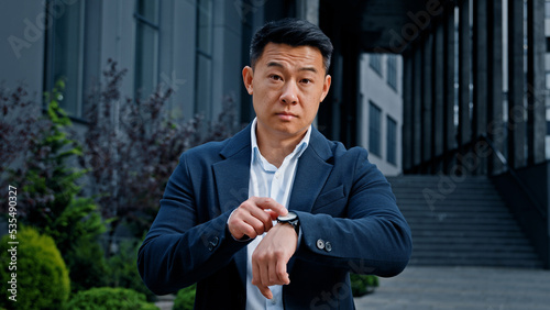 Angry worried 40s Asian businessman male frustrated boss manager CEO waiting meeting outdoors in city nervous annoyance frustrated pointing on wristwatch late time lateness deadline punctual wait