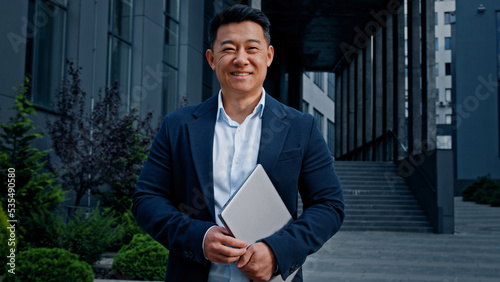Asian 40s middle-aged businessman entrepreneur employer man worker manager designer financial analyst standing in city near office company building in downtown posing holding laptop looking at camera