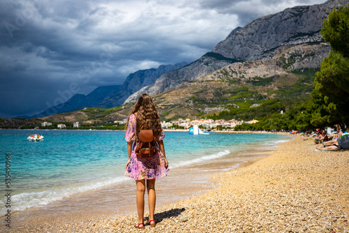 a beautiful woman in a purple dress with a leather backpack walks on a paradisiacal Croatian beach with mountains in the background, Makarska on the Adriatic coast © Jakub