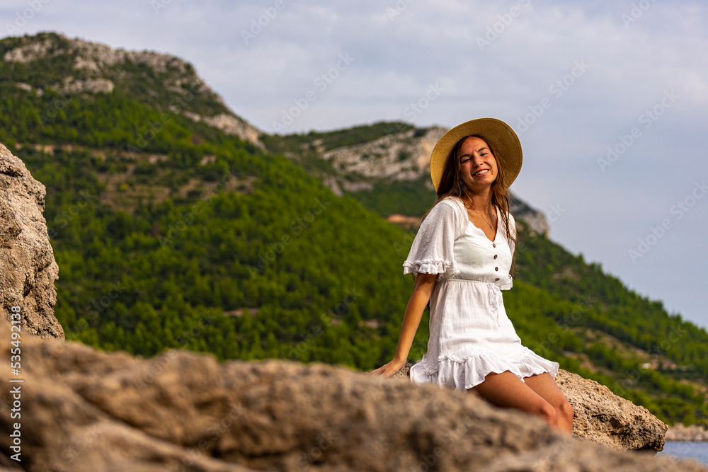 Naklejka premium beautiful tanned woman in white dress and hat rests on the rocks during sunset over paradise beach in croatia with green mountains in the background; peljesac peninsula 