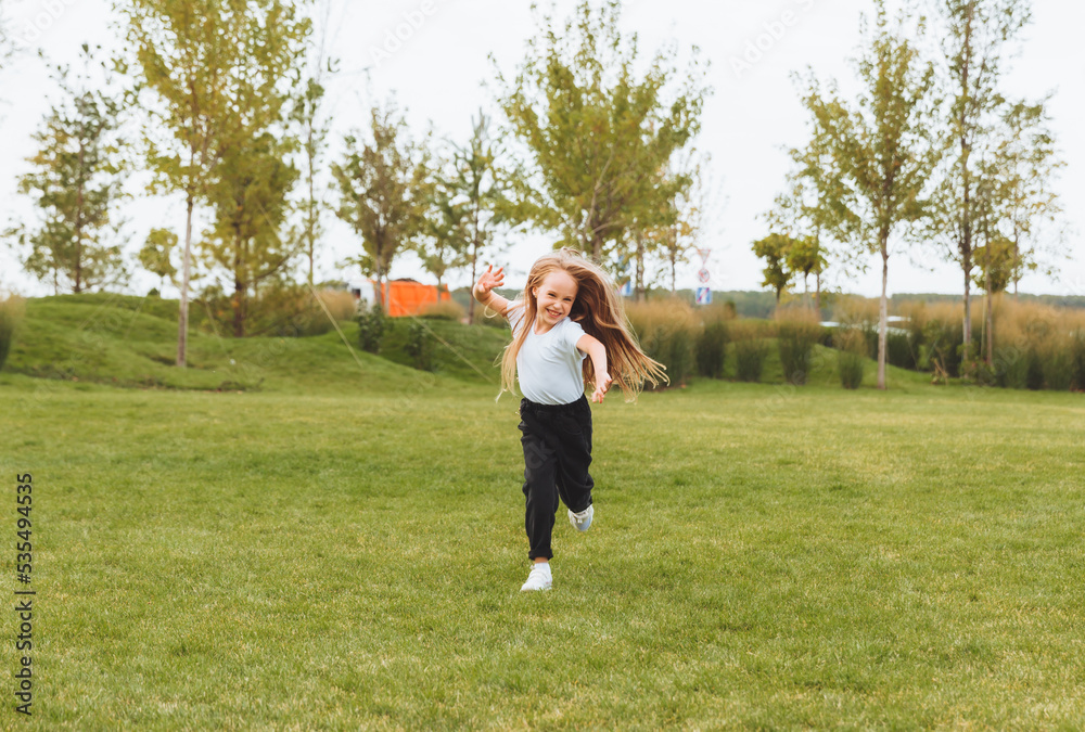a little cheerful girl with long hair runs through the grass in the park and rejoices.