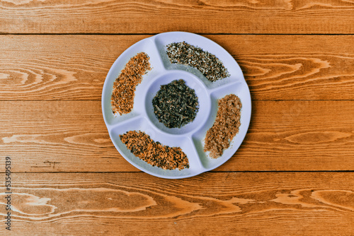 a large assortment of spices and herbs. it lies in a black plate. Peas, Pepper, salt, paprika, basil, turmeric. Indian, Georgian, Vietnamese cuisine. View from above.On a wooden board.Place for text	