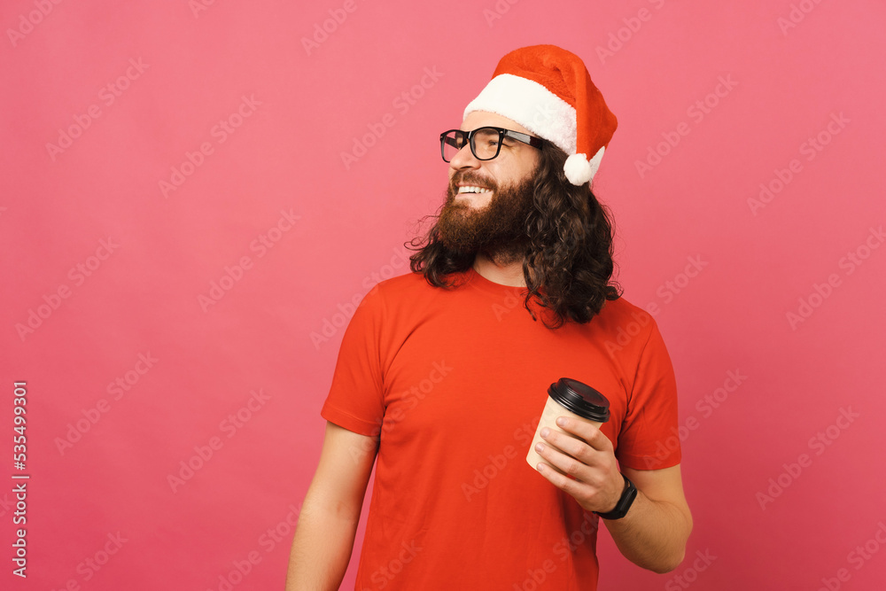 Photo of young man with beard and long hair holding cup of coffee to go and looking away at copyspace and wearing santa claus hat. Photo of young man with beard and long hair holding cup of coffee to go and looking away at copyspace and wearing santa claus hat.