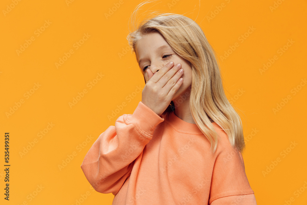 a beautiful girl, blonde, of school age, stands on an orange background in bright clothes and covers her mouth with her hand. Horizontal studio photography with blank space for advertising mockup a beautiful girl, blonde, of school age, stands on an orange background in bright clothes and covers her mouth with her hand. Horizontal studio photography with blank space for advertising mockup