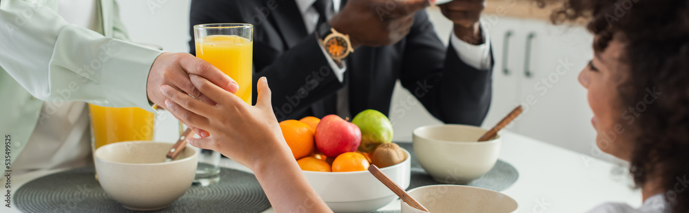 african american woman giving glass of orange juice to happy daughter near husband during breakfast, banner. african american woman giving glass of orange juice to happy daughter near husband during breakfast, banner.