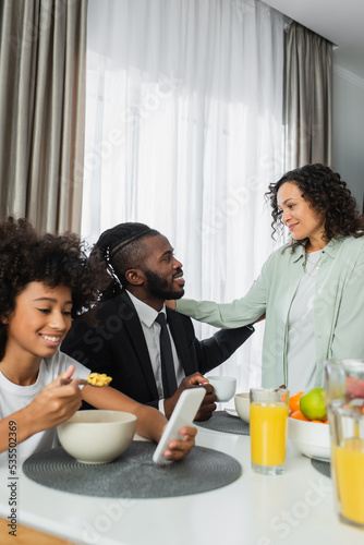 happy african american woman hugging husband near daughter using smartphone during breakfast.