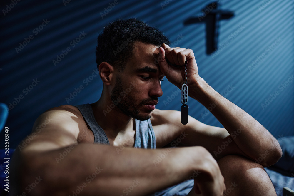 Obraz premium Young unhappy man with post traumatic syndrome holding silver chain with metallic medallion in hand touching his forehead