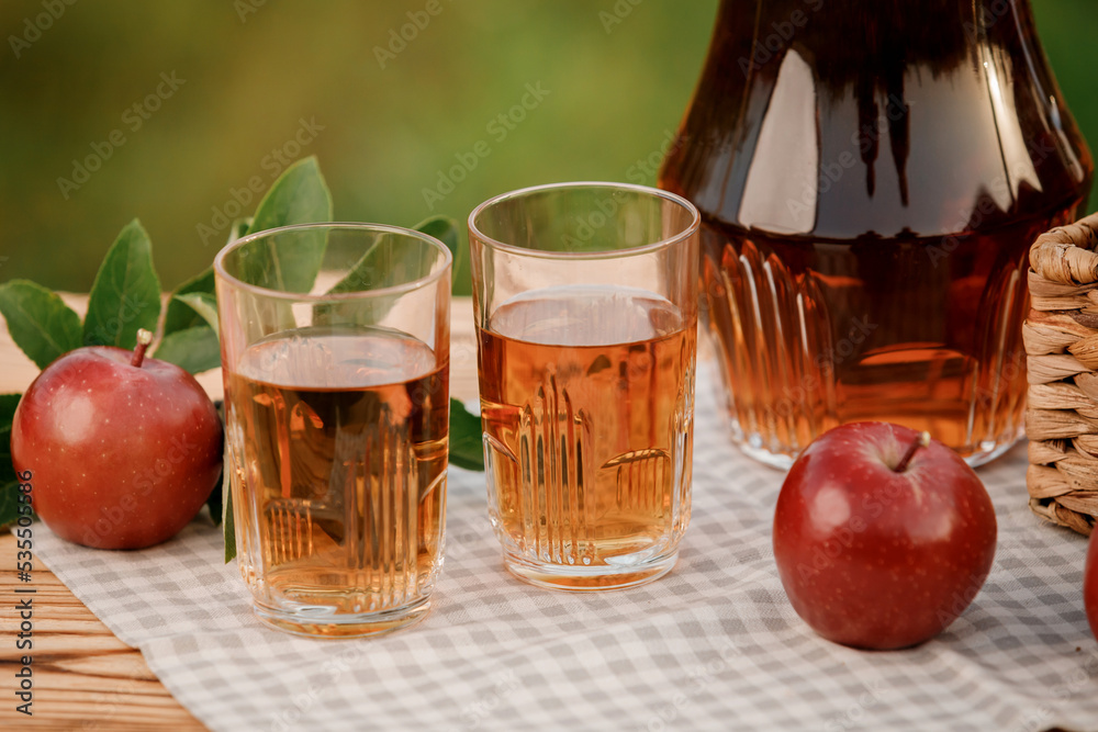 Two glasses with apple juice and basket with apples on wooden table with natural orchard background. Vegetarian fruit composition Two glasses with apple juice and basket with apples on wooden table with natural orchard background. Vegetarian fruit composition