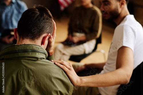 Rear view of young unhappy man suffering from loss or traumatic experience while one of attendants comforting him at session