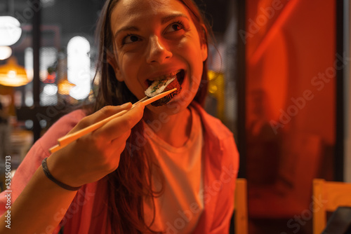 Portrait of smiling brunette woman eating sushi rolls with chopsticks and enjoying the taste. Red light. 