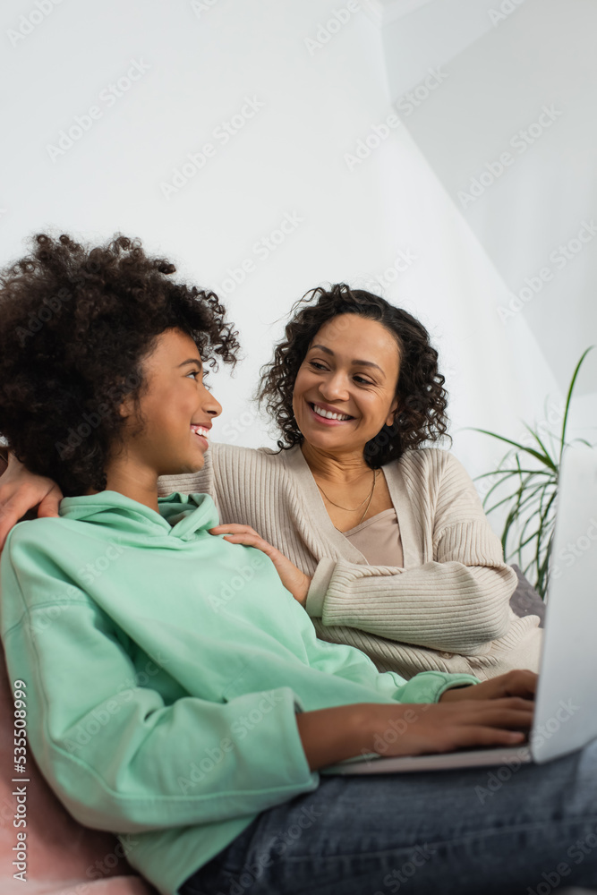 cheerful african american preteen girl using laptop near happy mother. cheerful african american preteen girl using laptop near happy mother.