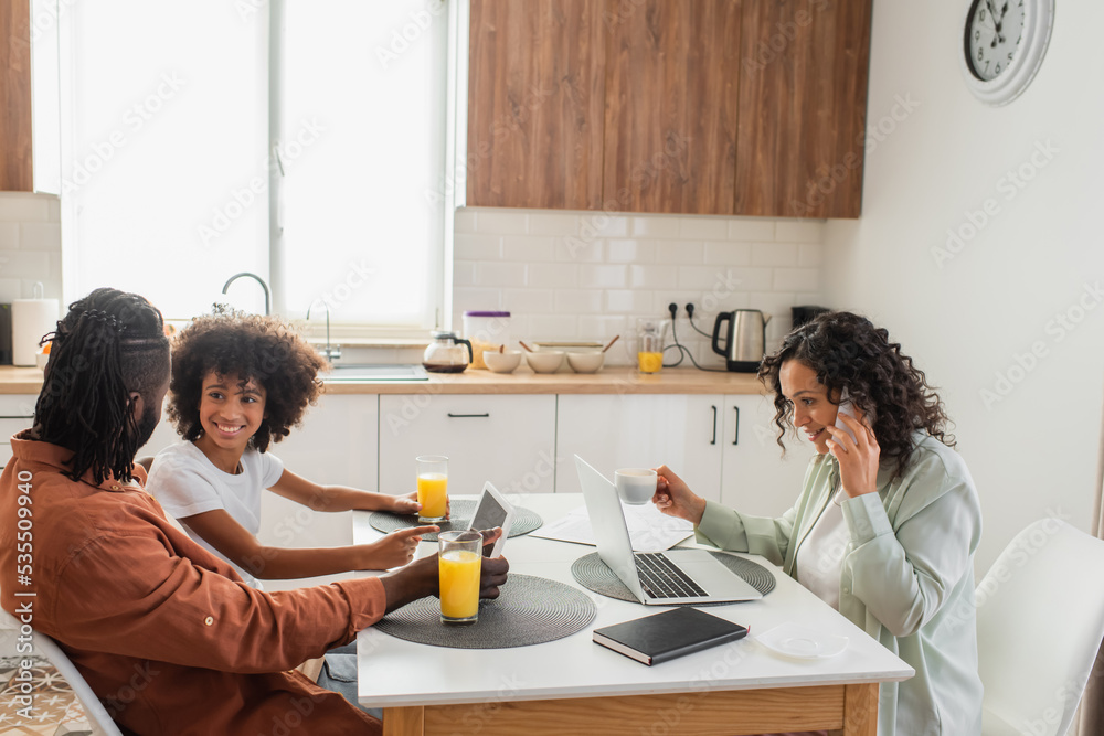 happy african american freelancer talking on smartphone near laptop while husband and daughter using digital tablet in kitchen.