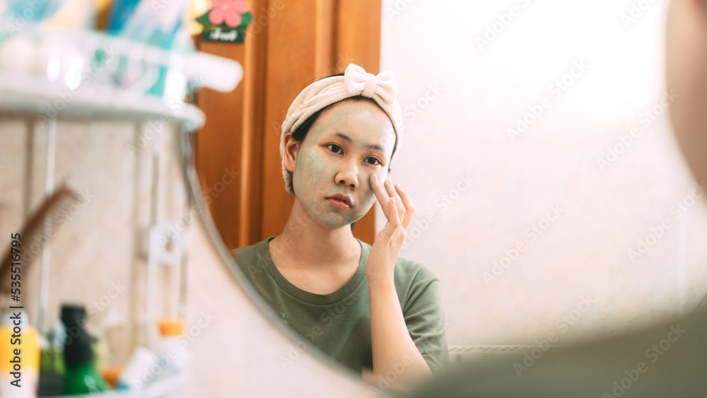 Young adult asian woman cleansing face with green clay facial mask for healing acne reflection with mirror in bathroom Young adult asian woman cleansing face with green clay facial mask for healing acne reflection with mirror in bathroom