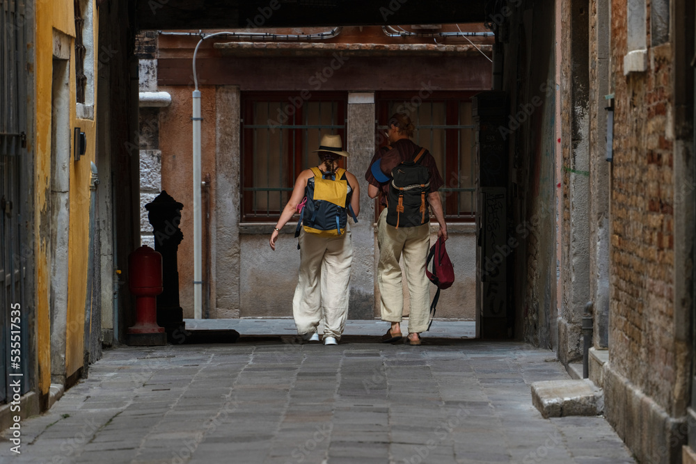 A couple of adult people with backpacks on their backs walk along a dark narrow passage in the city of Venice, Italy, historical landmarks tourist travel, Venetian streets