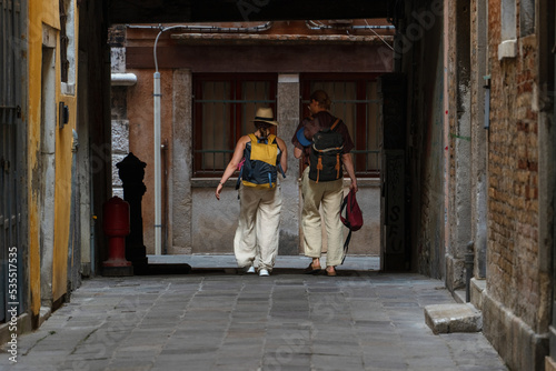 A couple of adult people with backpacks on their backs walk along a dark narrow passage in the city of Venice, Italy, historical landmarks tourist travel, Venetian streets