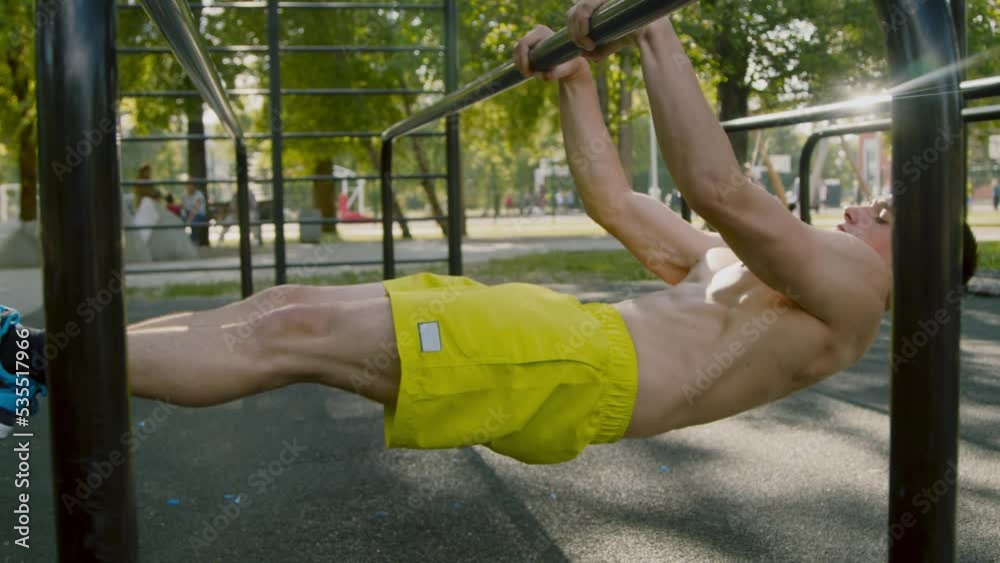 Modern strong young man wearing yellow shorts exercising in local city park doing front lever, slow motion shot