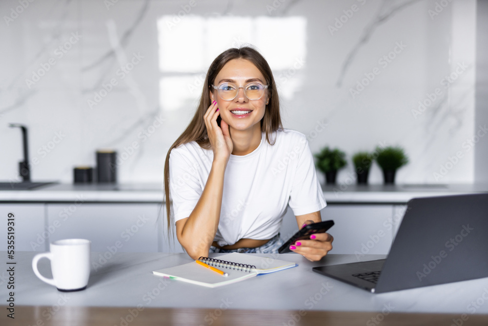 Busy multitask woman holds a smartphone with a shoulder and typing on the laptop keyboard. Freelance woman using laptop for remote work from home, sits at the kitchen desk and has phone conversation Busy multitask woman holds a smartphone with a shoulder and typing on the laptop keyboard. Freelance woman using laptop for remote work from home, sits at the kitchen desk and has phone conversation