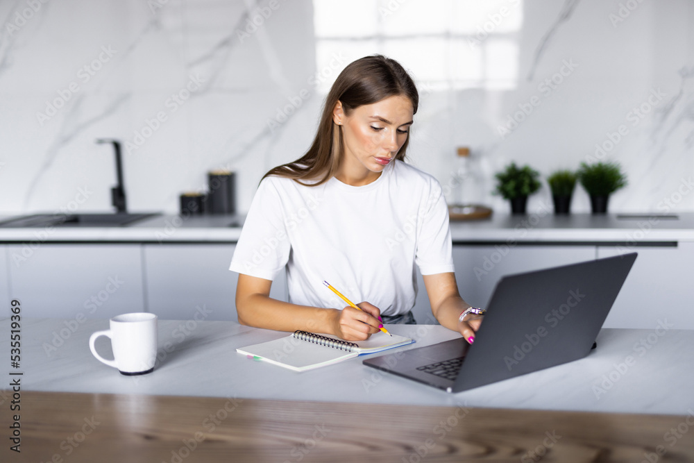Woman takes notes with pen, using laptop for remote work from home, write down some information sitting at the table. Multitasking concept