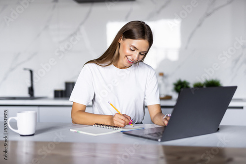 Woman takes notes with pen, using laptop for remote work from home, write down some information sitting at the table. Multitasking concept