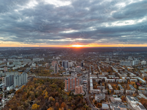 Aerial autumn city park sunset view with epic cloudscape. Botanical garden and Kharkiv city center. Residential district buildings and cars driving