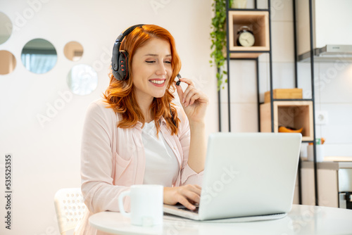 Young smiling friendly ginger girl working as agent at call center from home while drinking coffee. Wearing headset and typing on laptop.