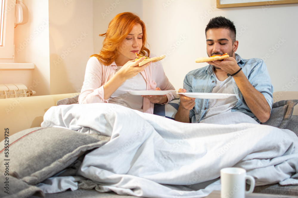 Cute young happy couple eating pizza together covered with blanket on sofa at home. Cute young happy couple eating pizza together covered with blanket on sofa at home.